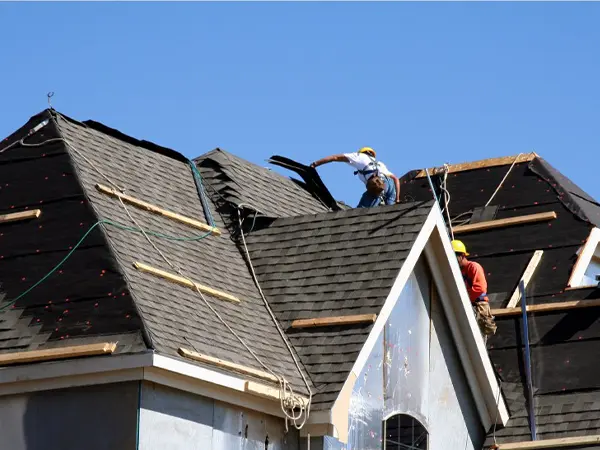 Workers replacing a damaged roof with new shingles under a clear blue sky.