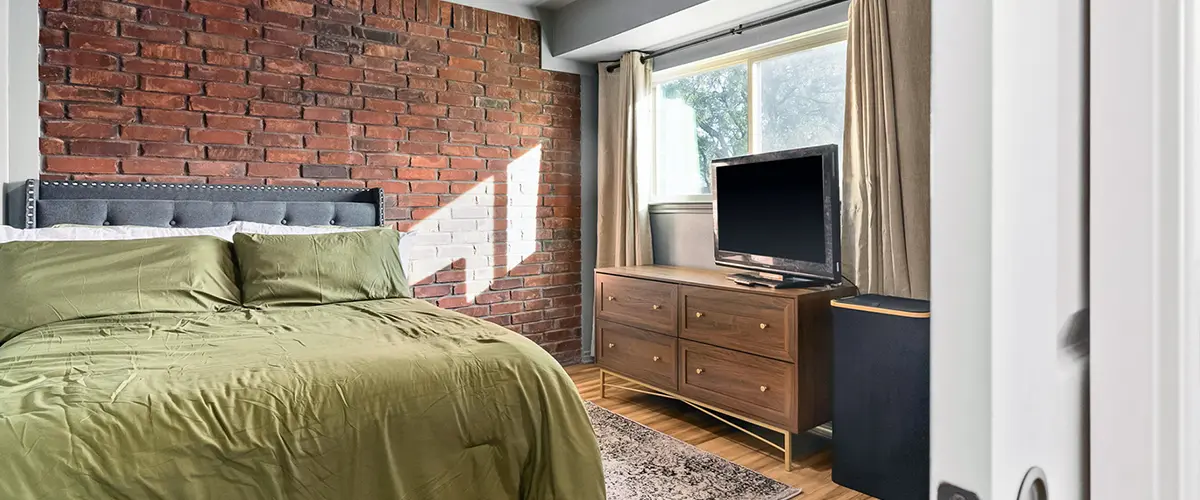 Stylish bedroom featuring an exposed brick accent wall, ceiling fan, green bedding, and a wooden dresser with a TV.