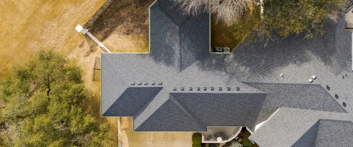 Aerial view of a large residential roof with dark shingles, multiple gables, and tree-lined surroundings.