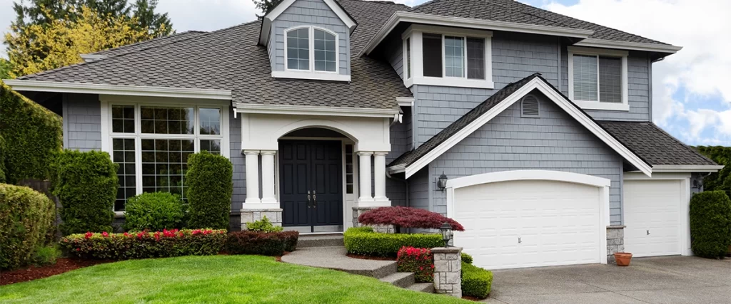A large, well-manicured suburban home with gray shingle siding, a dark gray front door, and prominent white-framed windows and dormers.