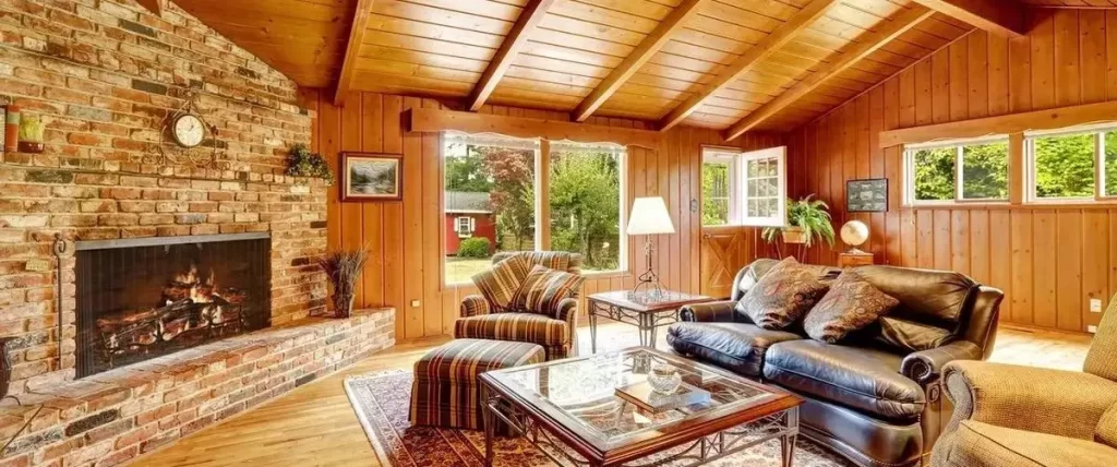 A cozy, rustic living room with wood paneled walls and an angled ceiling. The room features a large brick fireplace and multiple traditional-style windows looking out onto greenery.