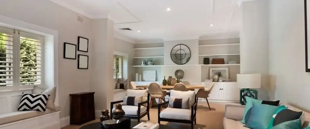 An open-concept living and dining space with built-in shelving, featuring a window seat with white plantation shutters and recessed lighting.