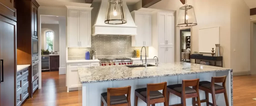 A luxury open-concept kitchen with white cabinetry, a large granite island, and a wide wooden range hood over a stainless steel range.