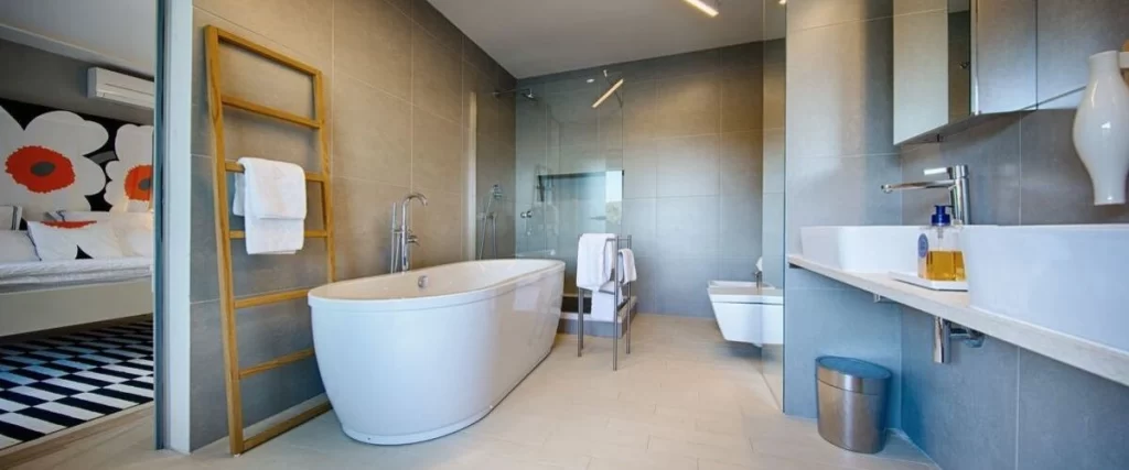 A minimalist modern bathroom with large gray wall tiles, featuring a white freestanding tub and a walk-in shower separated by a clear glass panel.