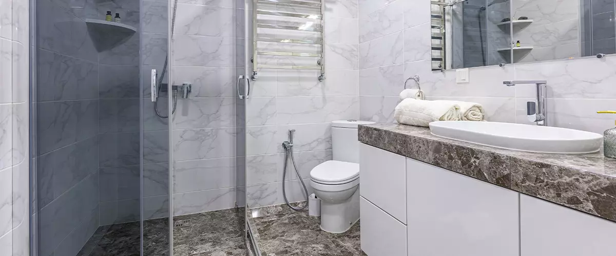 High-end bathroom featuring a glass walk-in shower with neutral stone wall and floor tiles, next to a light wood vanity with a marble countertop.