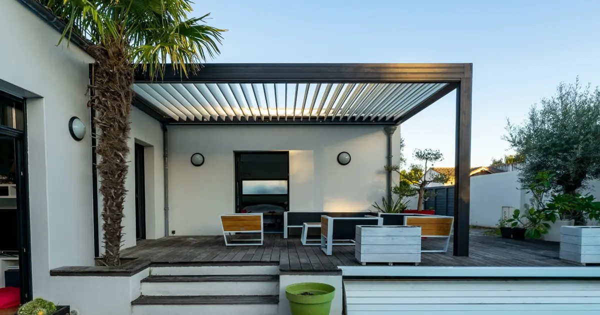 A modern black pergola with adjustable louvered roof slats extends from a white house over a wooden deck. Outdoor furniture is arranged on the deck, and a tall palm tree grows next to the pergola.