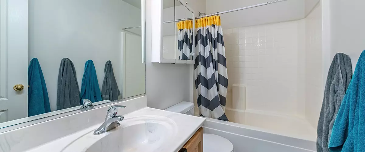 Standard bathroom with a white tub/shower unit, small vanity, mirror, medicine cabinet, and a grey and yellow chevron shower curtain.