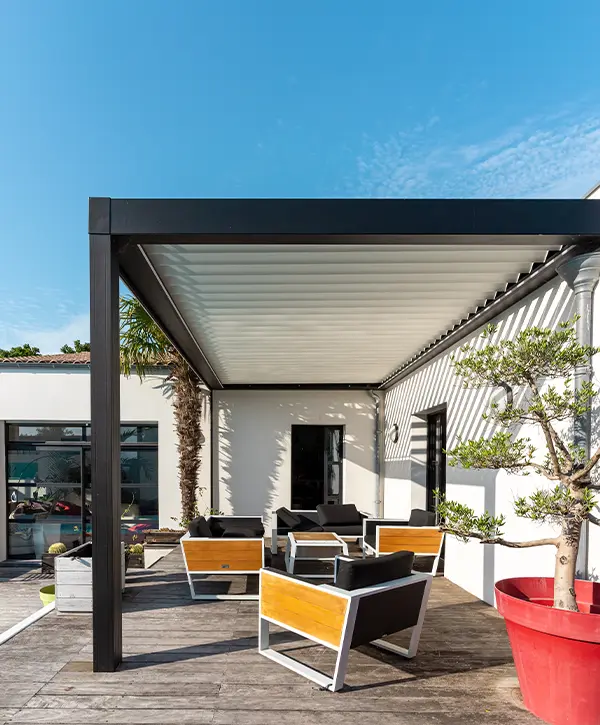 Sleek black-framed pergola with white adjustable louvered slats attached to a modern white home, covering an outdoor seating area.
