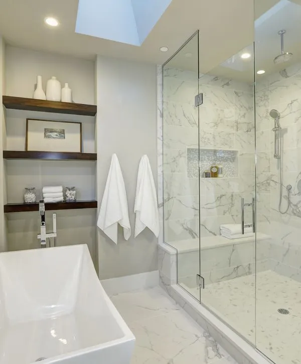 Modern bathroom conversion with white marble tile, a glass walk-in shower with a seat, and a deep white freestanding tub.