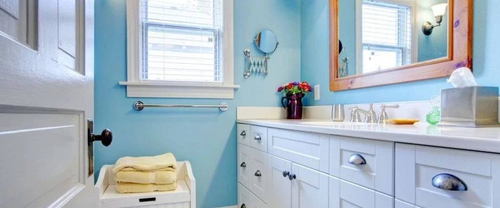 Cheerful bathroom with light blue walls, a white vanity cabinet and countertop, a window with blinds, and yellow towels stacked on a laundry hamper.
