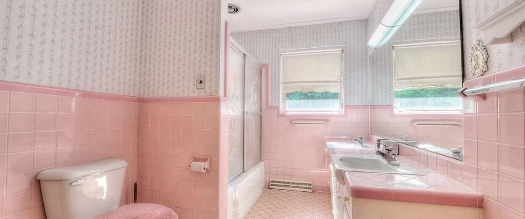 Retro bathroom design featuring bright pink square tiles covering the lower walls, pink trim, and patterned wallpaper above, with a toilet, tub, and vanity.