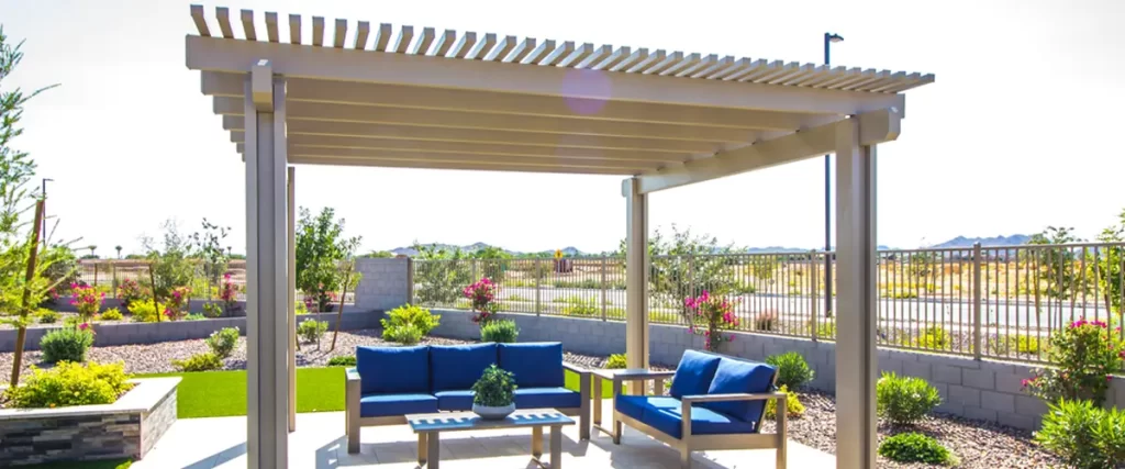 A light-colored metal pergola provides shade over blue outdoor sofas and a coffee table in a modern desert landscape with gravel, manicured bushes, and a distant fence.