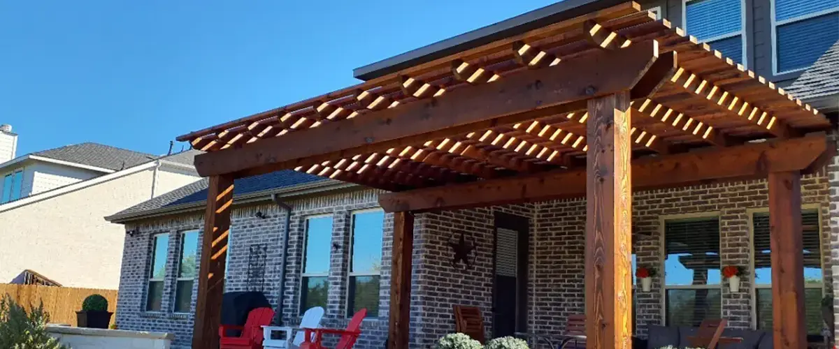 A custom dark-stained cedar wood pergola structure attached to the rear of a brick home, providing shade over a patio area with red and white outdoor chairs.