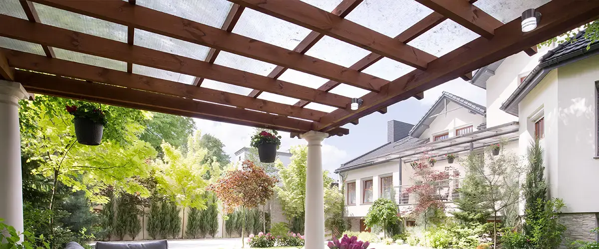 A dark wood pergola with a transparent protective roof cover and hanging flower pots overlooking a lush green garden and modern house.