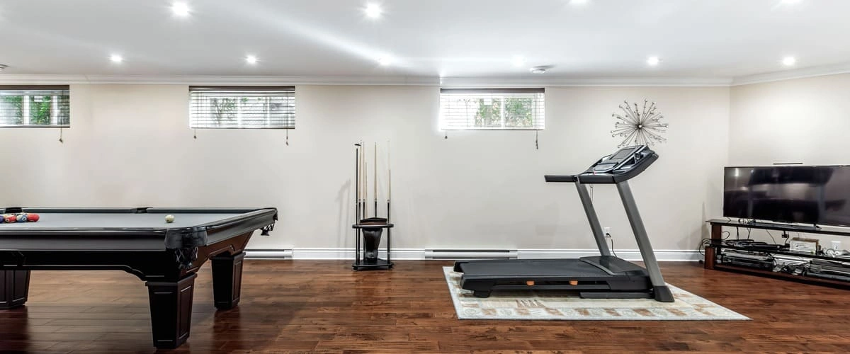 A spacious finished garage converted into a multi-purpose recreation room featuring dark wood laminate flooring, a professional pool table, and a treadmill setup for a home gym.