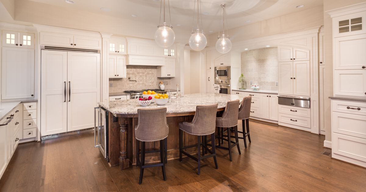 A spacious, luxury white kitchen featuring a large granite island with seating, designed for aging in place remodeling with wide clearance for mobility and bright pendant lighting.