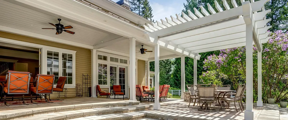 A large white attached pergola providing shade over a stone patio featuring orange cushioned lounge chairs, outdoor ceiling fans, and a dining set.
