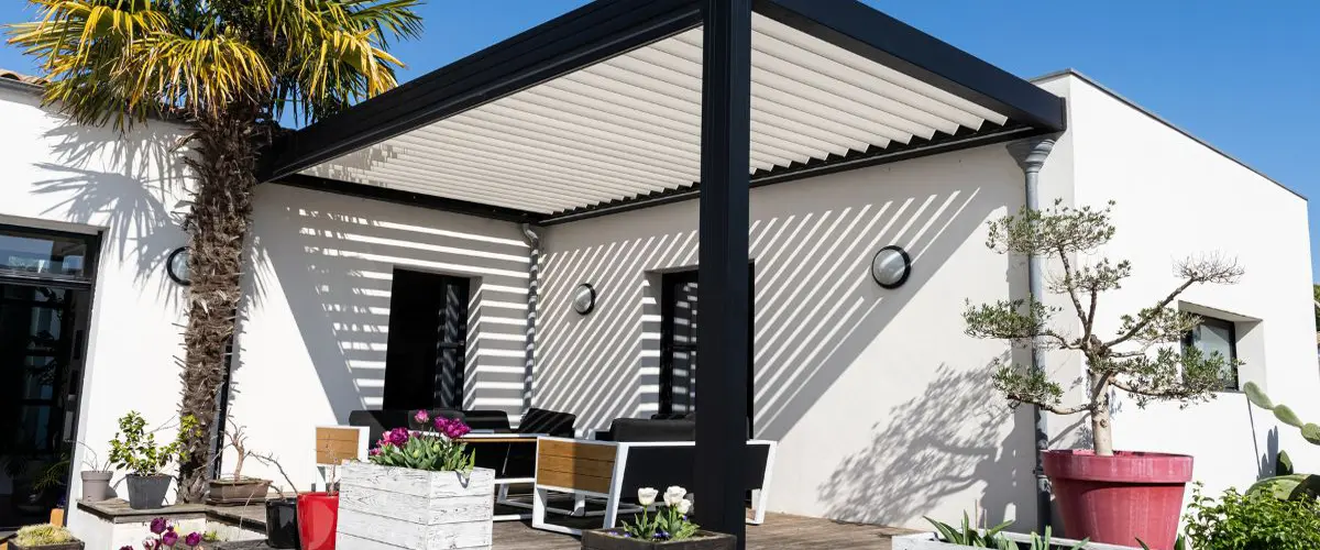A contemporary black aluminum bioclimatic pergola with an adjustable white louvered roof installed over a modern patio with outdoor furniture and potted plants.