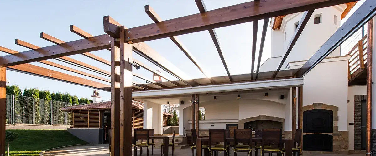 A large modern freestanding wooden pergola with a retractable canopy covering an outdoor dining table and luxury stone summer kitchen area.