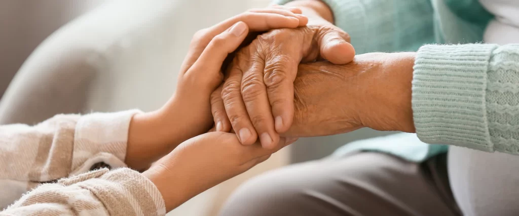 A close-up of a younger person’s hands gently holding the weathered hands of a senior, showing emotional support for aging in place.