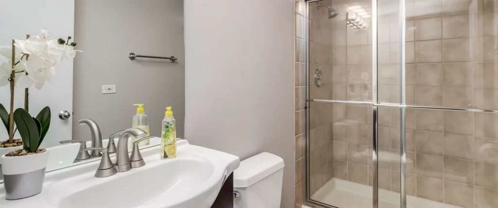A clean minimalist bathroom showing a white pedestal sink with chrome faucets and a sliding glass shower door with tan tiles.