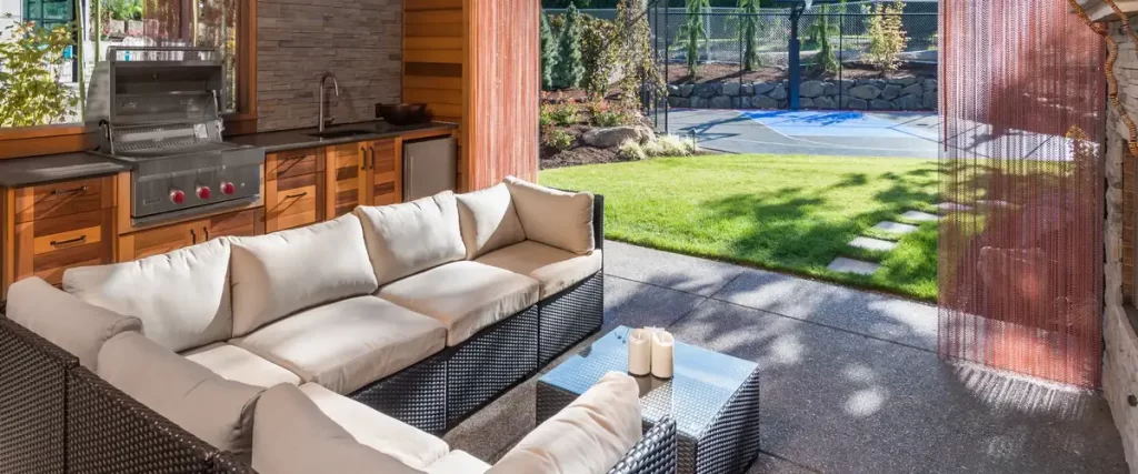 A covered outdoor living area with a large sectional sofa, glass coffee table, and a built-in outdoor grill station.