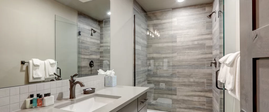 Modern bathroom interior showing a gray vanity with a bronze faucet next to a glass-enclosed shower featuring wood-look porcelain wall tiles.