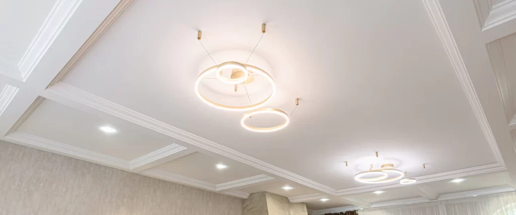 A white recessed coffered ceiling decorated with several modern minimalist gold circular ring pendant lights and integrated spotlights.