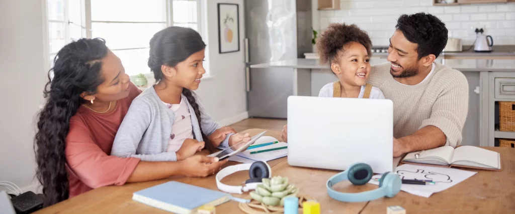 A smiling family sitting together at a wooden dining room table using a laptop and tablet in a bright home.