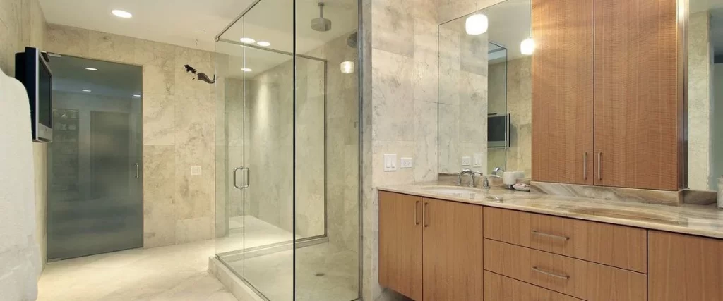 High-end master bathroom featuring custom wood cabinetry, marble countertops, and a large seamless glass shower enclosure with neutral stone tiling.