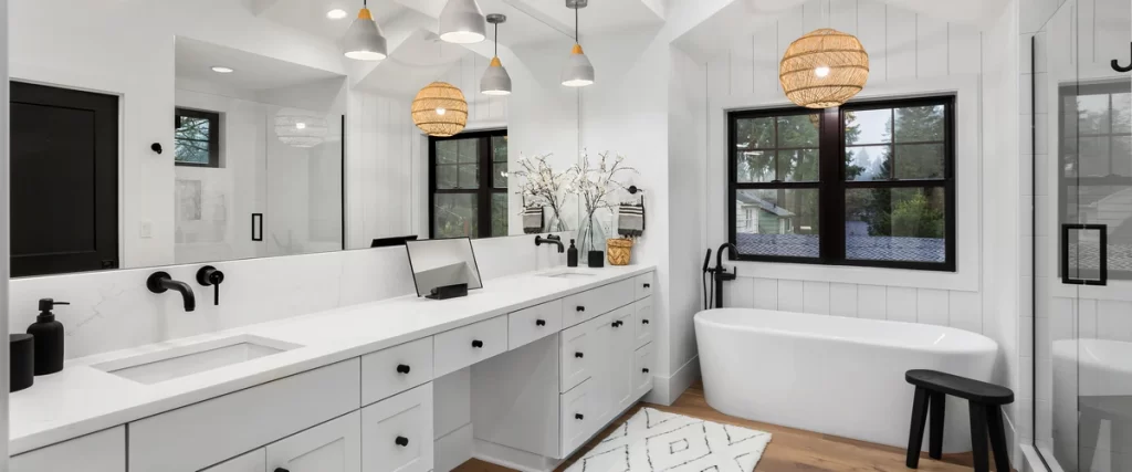 A spacious white master suite featuring a long double vanity, a soaking tub, and large black-framed windows looking out to a forest view.