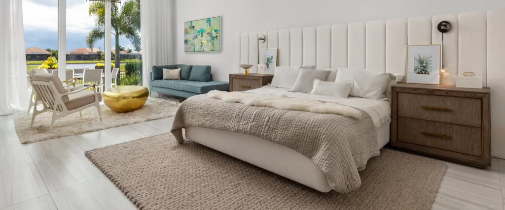 A luxurious primary suit bedroom featuring a large bed, neutral tones, a blue sofa, and large windows overlooking a lake.