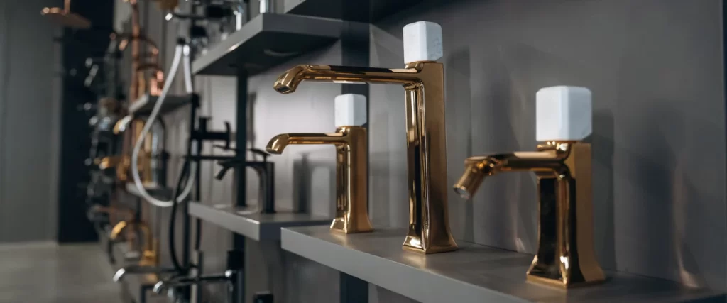 A row of premium polished gold bathroom faucets with white marble handles displayed on dark grey shelves in a showroom setting.