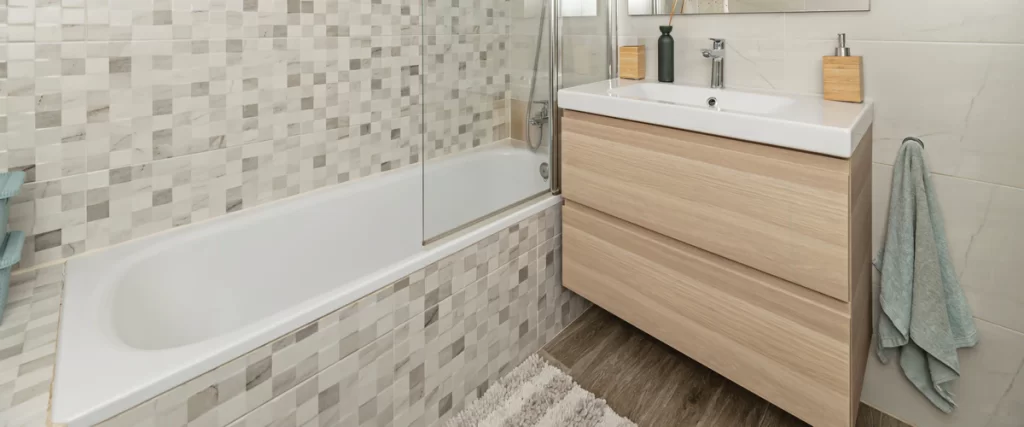 A light wood bathroom vanity featuring deep built-in storage drawers located next to a white bathtub with grey mosaic tile accents.