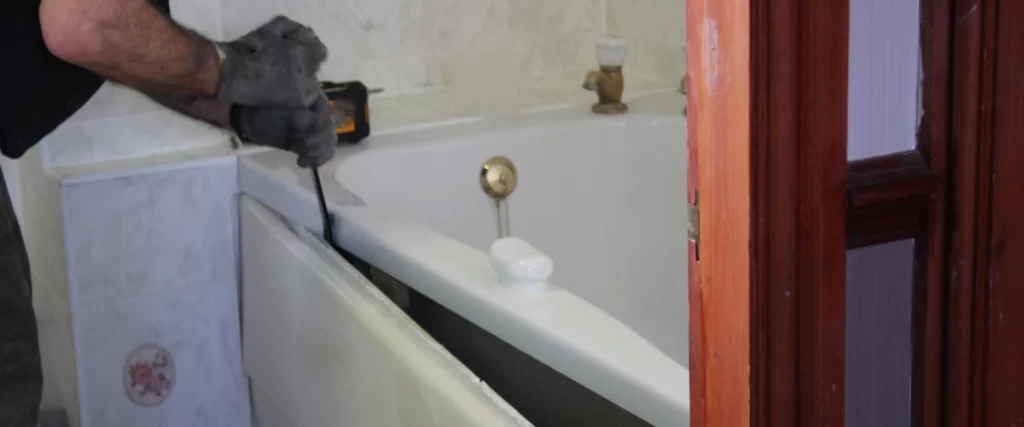 A close-up of a worker using a pry bar to remove a side panel from an old white bathtub during a shower conversion project.
