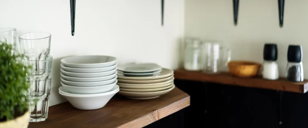 Wooden open shelving in a kitchen holding stacked white ceramic bowls, glass tumblers, and seasoning jars.