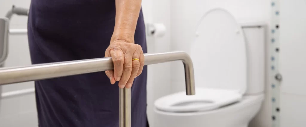An elderly woman holding onto a stainless steel safety grab bar next to a toilet to maintain balance while aging in place.
