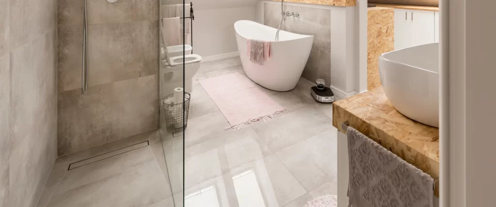 A wide view of a luxury bathroom floor and walls covered in high-gloss light grey large tiles featuring a freestanding soaking tub and a walk-in glass shower.