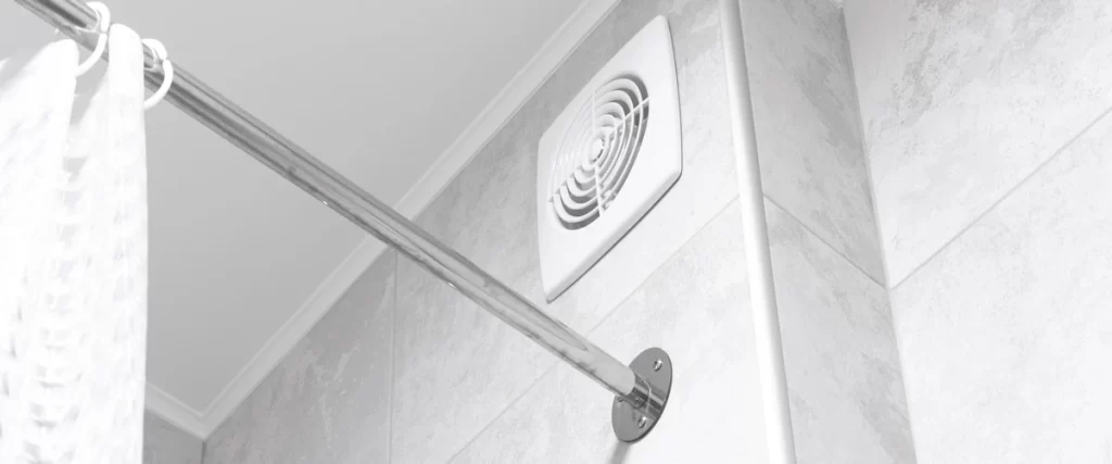 A close-up shot of a square white plastic ventilation fan installed high on a grey marble-patterned tiled wall near a chrome shower rod.