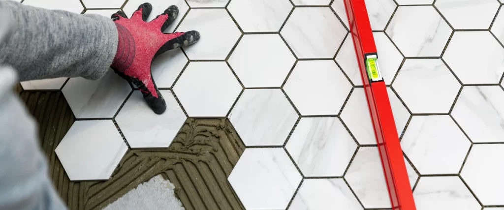 A close-up view of a professional bathroom tile installation where a gloved hand is laying white hexagonal marble tiles into thinset mortar next to a red spirit level.