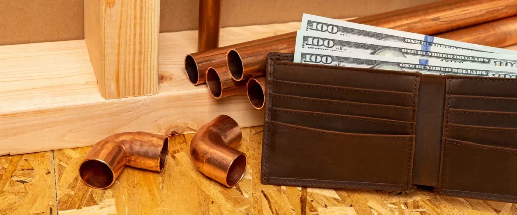 A leather wallet filled with hundred-dollar bills sitting next to copper plumbing pipes, representing a bathroom remodeling budget.