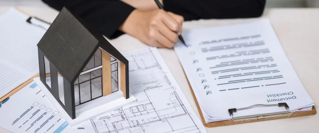 Close-up of a person marking a document checklist for a bathroom remodeling contract next to a scale house model and architectural drawings.