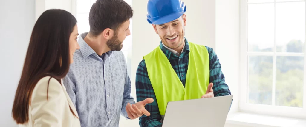 A smiling bathroom remodeling contractor in a blue hard hat and safety vest using a laptop to maintain clear communication with a couple.