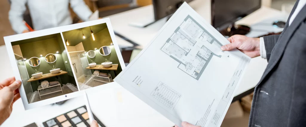 A person holding a professional bathroom remodeling plan blueprint next to photos of a finished green bathroom with double vanity sinks and round mirrors.