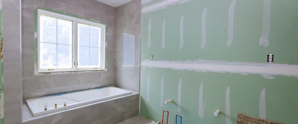 Progress shot of the bathroom remodeling process featuring green moisture-resistant drywall, plumbing stubs for a vanity, and a newly installed bathtub with grey marble tile surround.