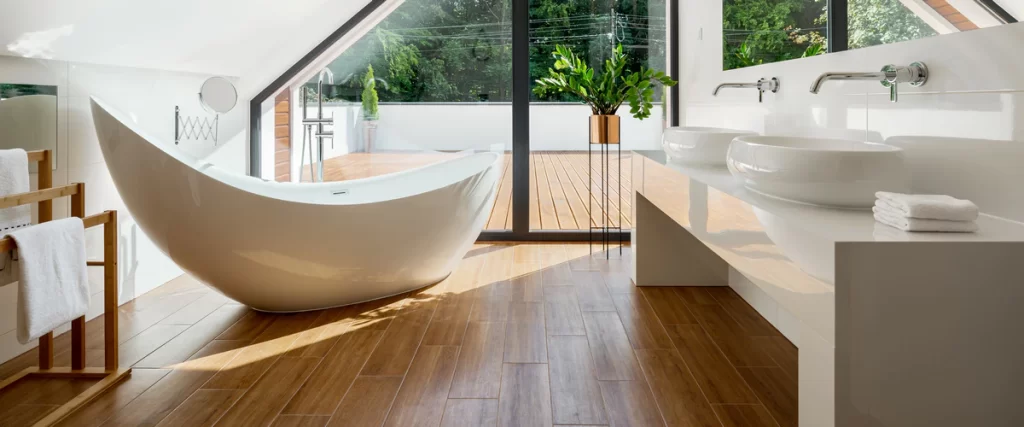 A sunlit attic bathroom featuring wood-look tile flooring, a unique curved white bathtub, and double vessel sinks facing a large glass door leading to a wooden deck.