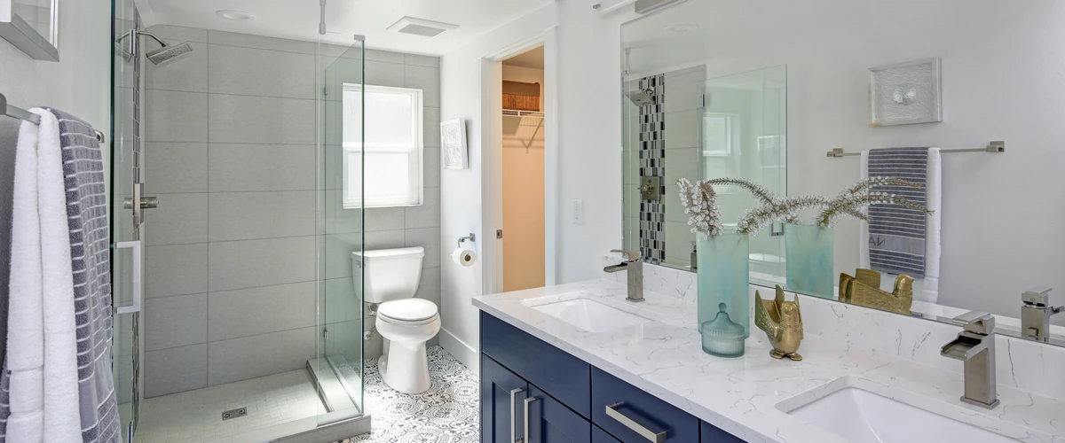 A bright bathroom featuring a navy blue double vanity, patterned floor tiles, and a glass-enclosed shower with large format grey wall tiles.