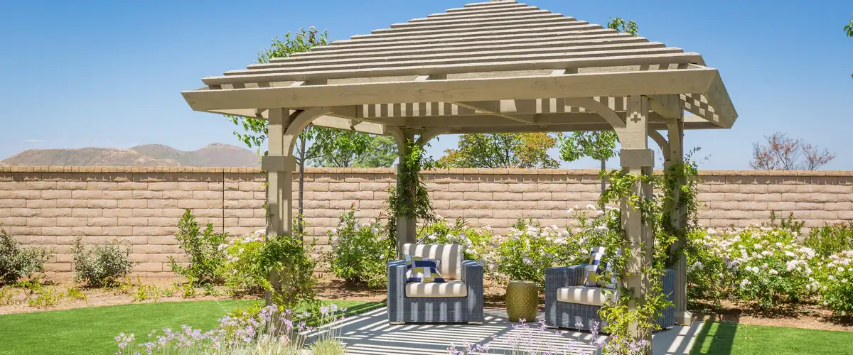 A freestanding wooden garden pergola with a unique tiered slatted roof design and climbing greenery, providing shade for a cozy outdoor seating area with striped chairs.