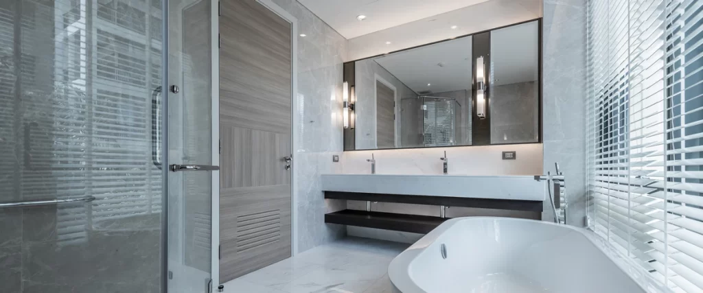 Luxury bathroom design with grey marble tiles, a wide double vanity, modern wall-mounted light sconces, and a large white bathtub near a window with blinds.
