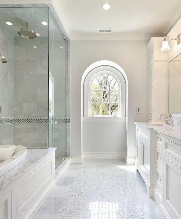 A bright, luxurious bathroom conversion, Salado, TX featuring a white marble walk-in glass shower, a built-in tub, dual white vanities, and a distinct arched window.